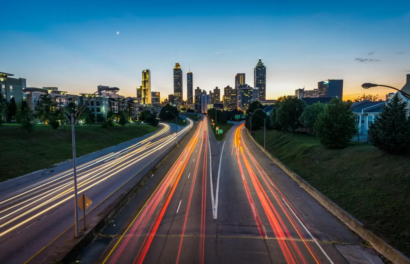 A long-exposure photograph of a city skyline at dusk, with bright light trails from cars on two highways leading toward the illuminated buildings. The image represents the fast-paced and dynamic urban real estate market, highlighting a streamlined path for homeowners.