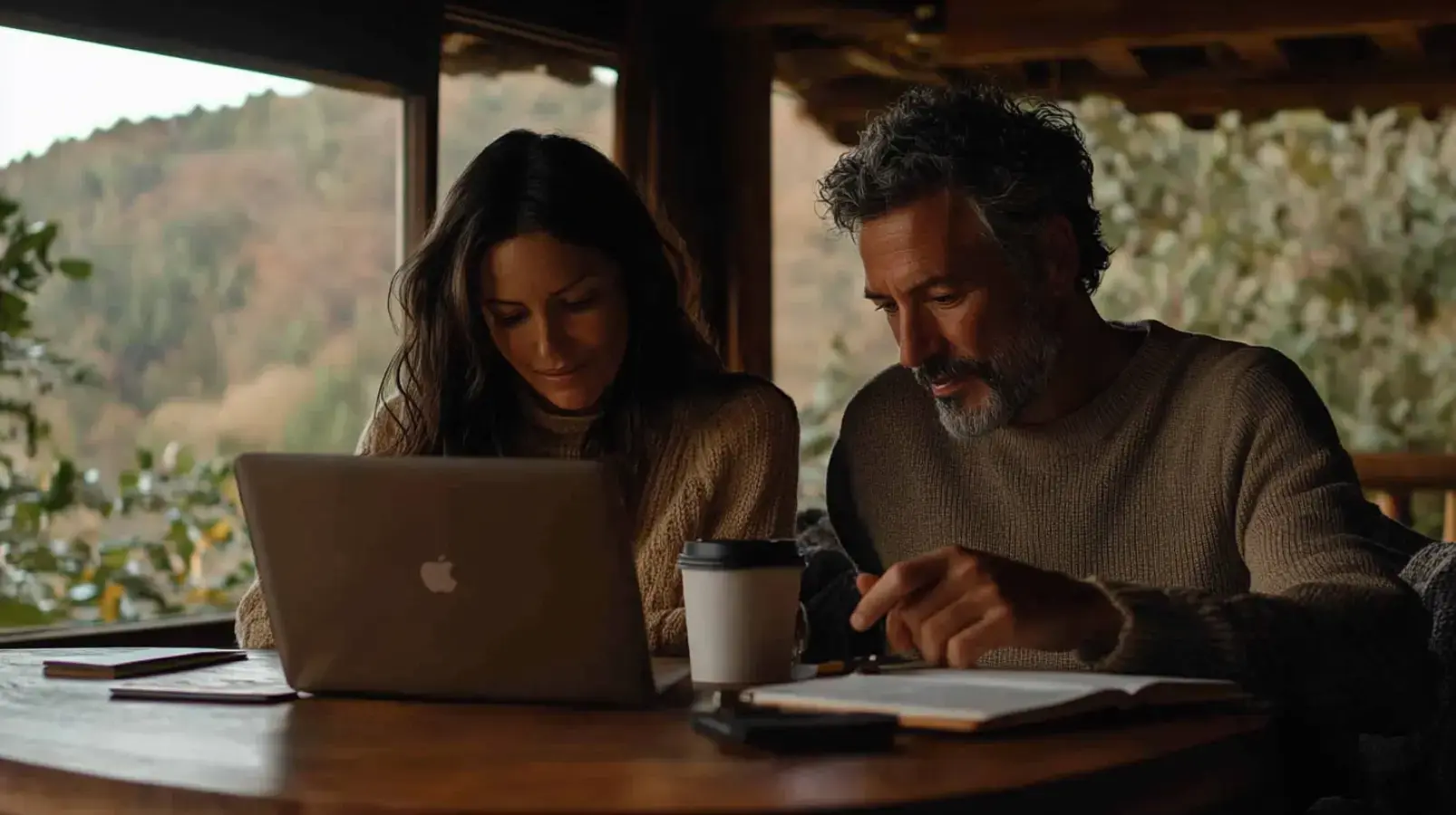 A middle-aged couple sits together at a table, intently reviewing documents and a laptop with a scenic, mountainous view behind them. The image represents a couple collaboratively engaging in serious financial planning, symbolizing the thoughtful process of a sale-leaseback.