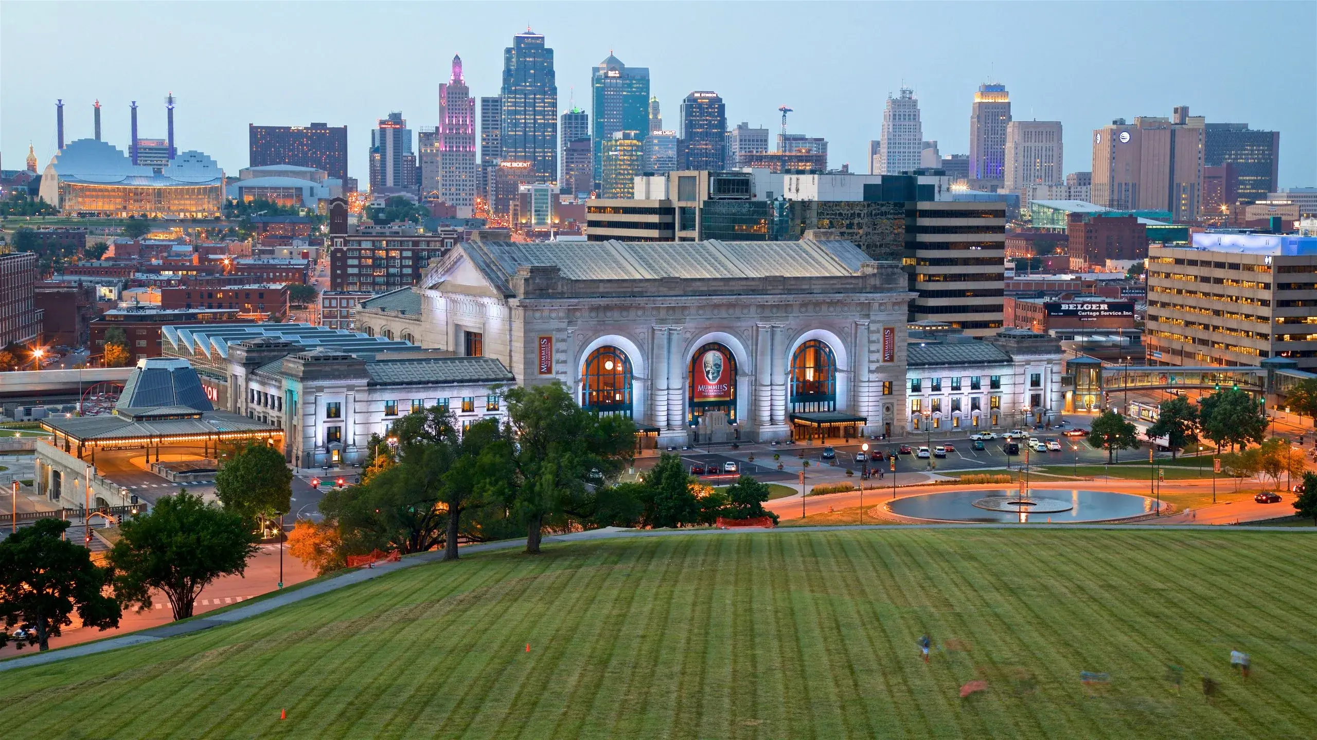 The Kansas City, Missouri skyline at dusk, with Union Station and a large green lawn in the foreground.