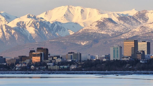 The Anchorage, Alaska city skyline with a backdrop of large, snow-capped mountains and a frozen body of water in the foreground.