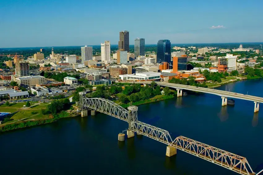 An aerial view of the Little Rock, Arkansas skyline on a sunny day, with two bridges crossing the Arkansas River.
