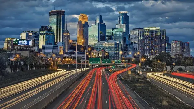 The Minneapolis, Minnesota skyline at night, with long exposure light trails from traffic on a busy highway in the foreground.